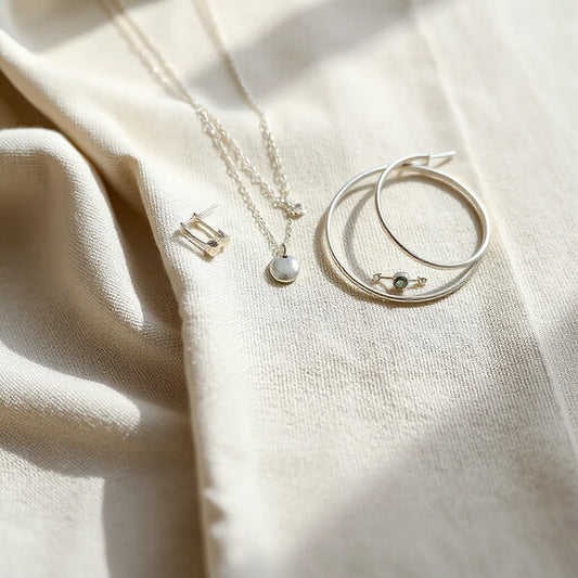 Layered sterling silver necklaces, small hoops, and bangles arranged on neutral fabric