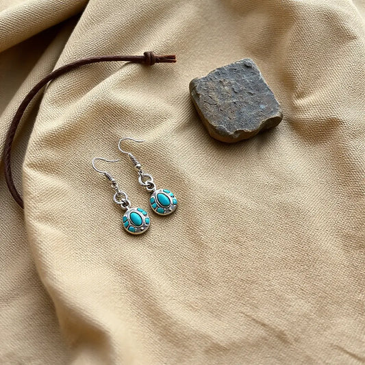 Silver-toned western inspired earrings with concho and turquoise-inspired accents on warm neutral fabric